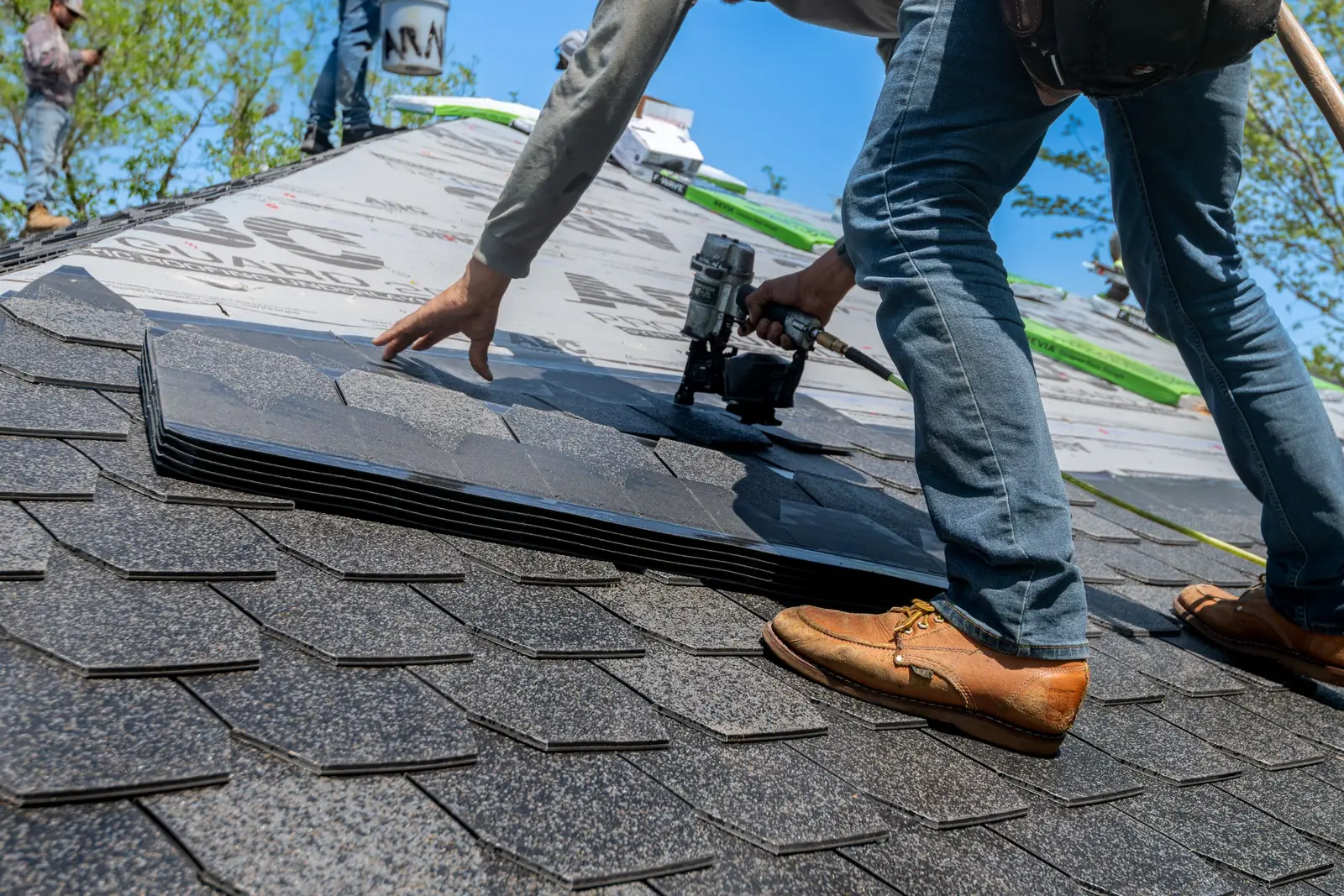 A roofer on a pitched roof fixing a row of new asphalt shingles with a nail gun